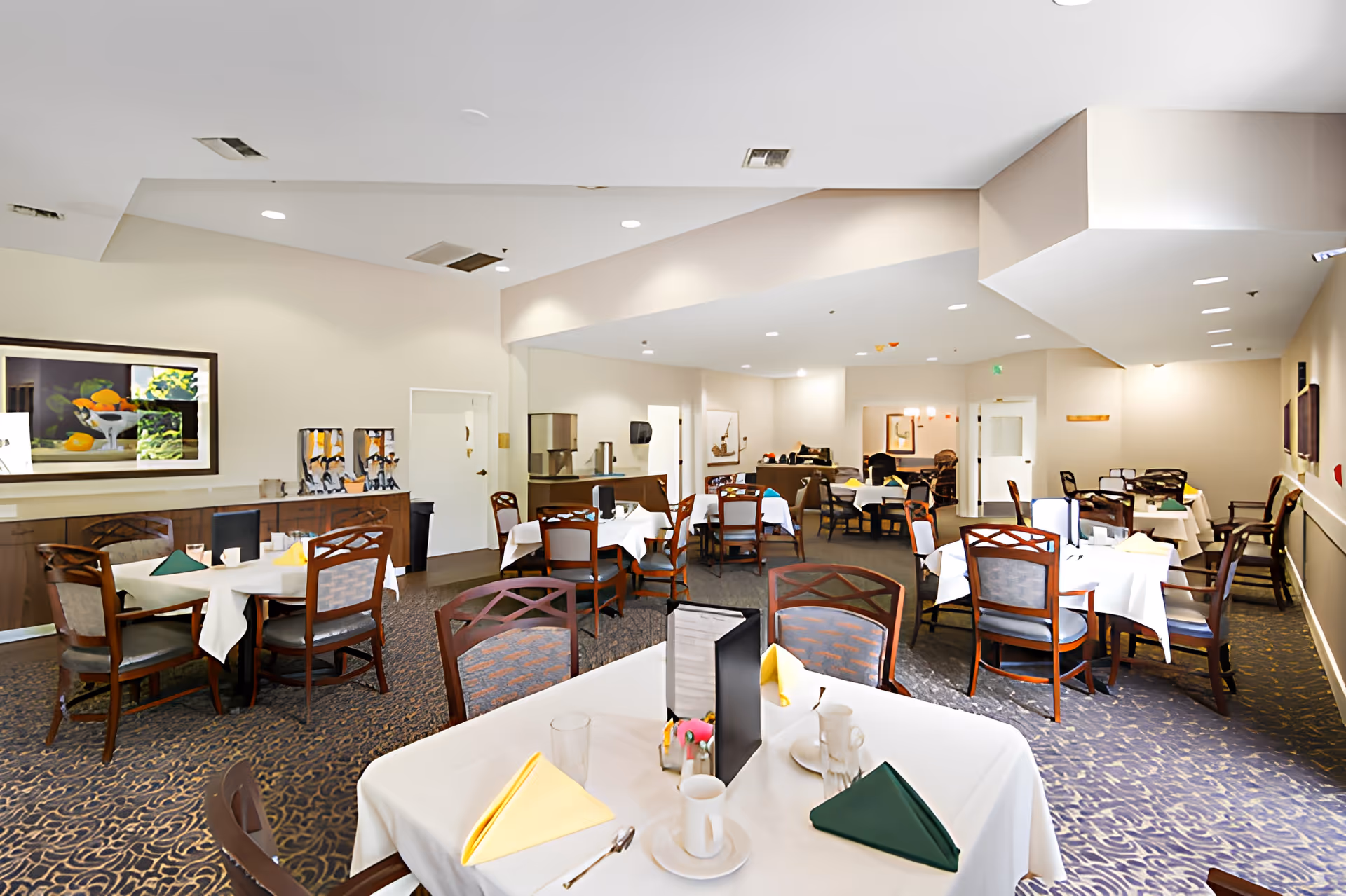 Spacious dining room with multiple tables covered in white linens, place settings and wooden chairs, plus a buffet/service area.
