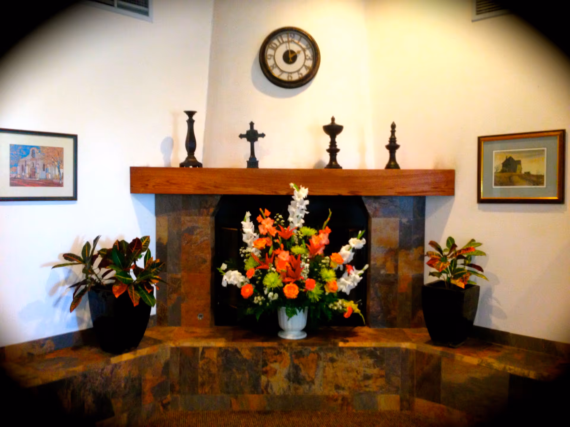 Decorative fireplace mantel with a clock and candlesticks above a large floral arrangement, flanked by potted plants and framed pictures.