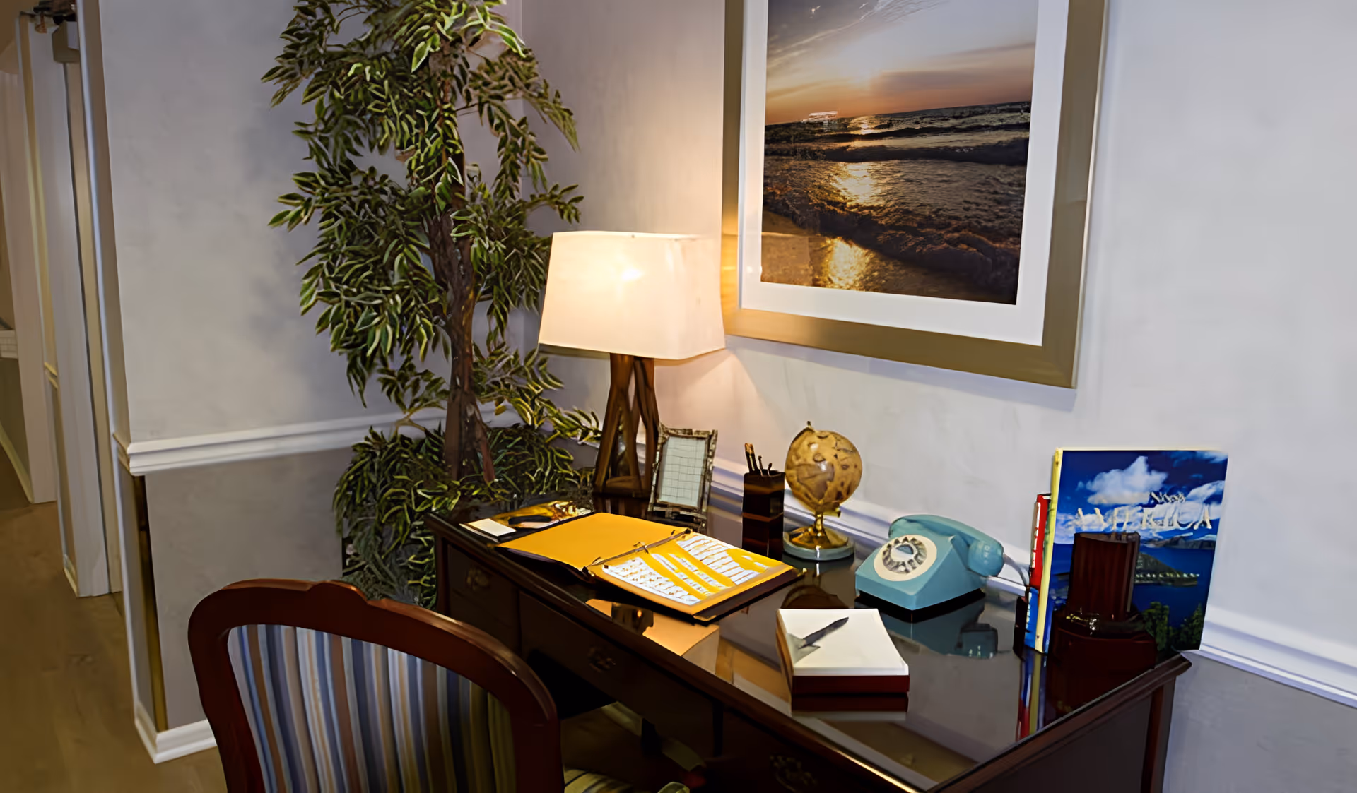 A cozy office corner with a wooden desk and a striped chair. On the desk are a table lamp, a small globe, a vintage rotary phone, an open yellow folder, a pen holder, a tissue box, and some books. A large framed photograph of a sunset over the ocean hangs on the wall above the desk. A tall leafy plant is positioned next to the desk.