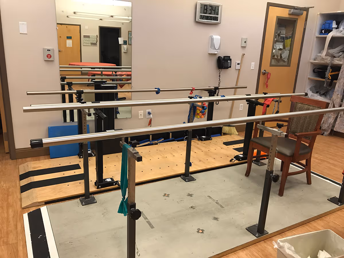 An indoor physical therapy room with parallel bars for walking exercises, a wooden chair, a wall-mounted clock showing 8:34, a wall phone, a large mirror, and shelves with various supplies.