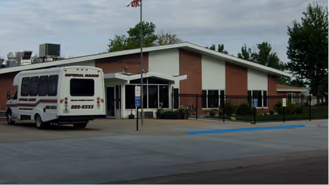 Exterior view of Imperial Manor Parkview Heights building with a white shuttle bus parked in front. The building is single-story with brick walls and a white entrance canopy. There is a flagpole with an American flag near the entrance and a black metal fence surrounding part of the property. The sky is cloudy.