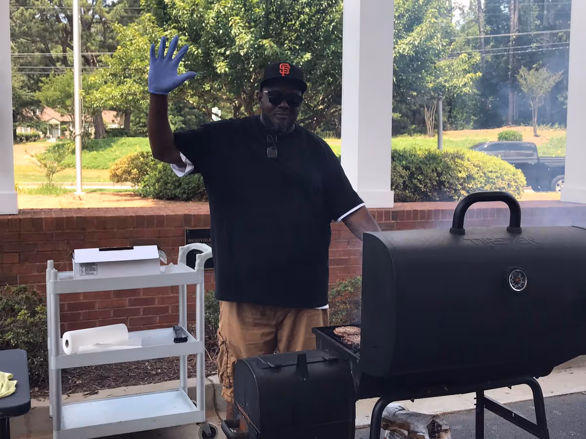 A man wearing a black t-shirt, tan shorts, a black San Francisco Giants cap, sunglasses, and a blue glove on one hand is standing outdoors next to a black smoker grill with smoke coming out. He is waving with his gloved hand. Behind him is a white cart with paper towels and a box, a brick wall, and green bushes and trees in the background.