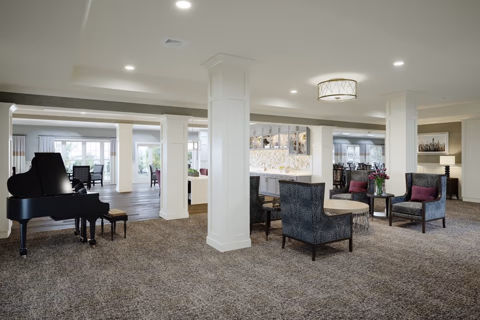A spacious and well-lit common area in a senior living facility featuring a black grand piano with a matching bench on the left, several comfortable armchairs with purple cushions arranged around small round tables, white columns, and a kitchenette with white cabinetry and a decorative backsplash in the background. Large windows and glass doors allow natural light to fill the room.