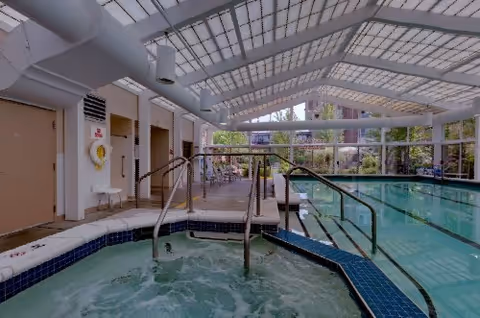 Indoor swimming pool and adjacent hot tub under a white skylight roof with glass walls.
