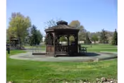 A wooden gazebo situated in the middle of a well-maintained grassy area with trees and a clear blue sky in the background.