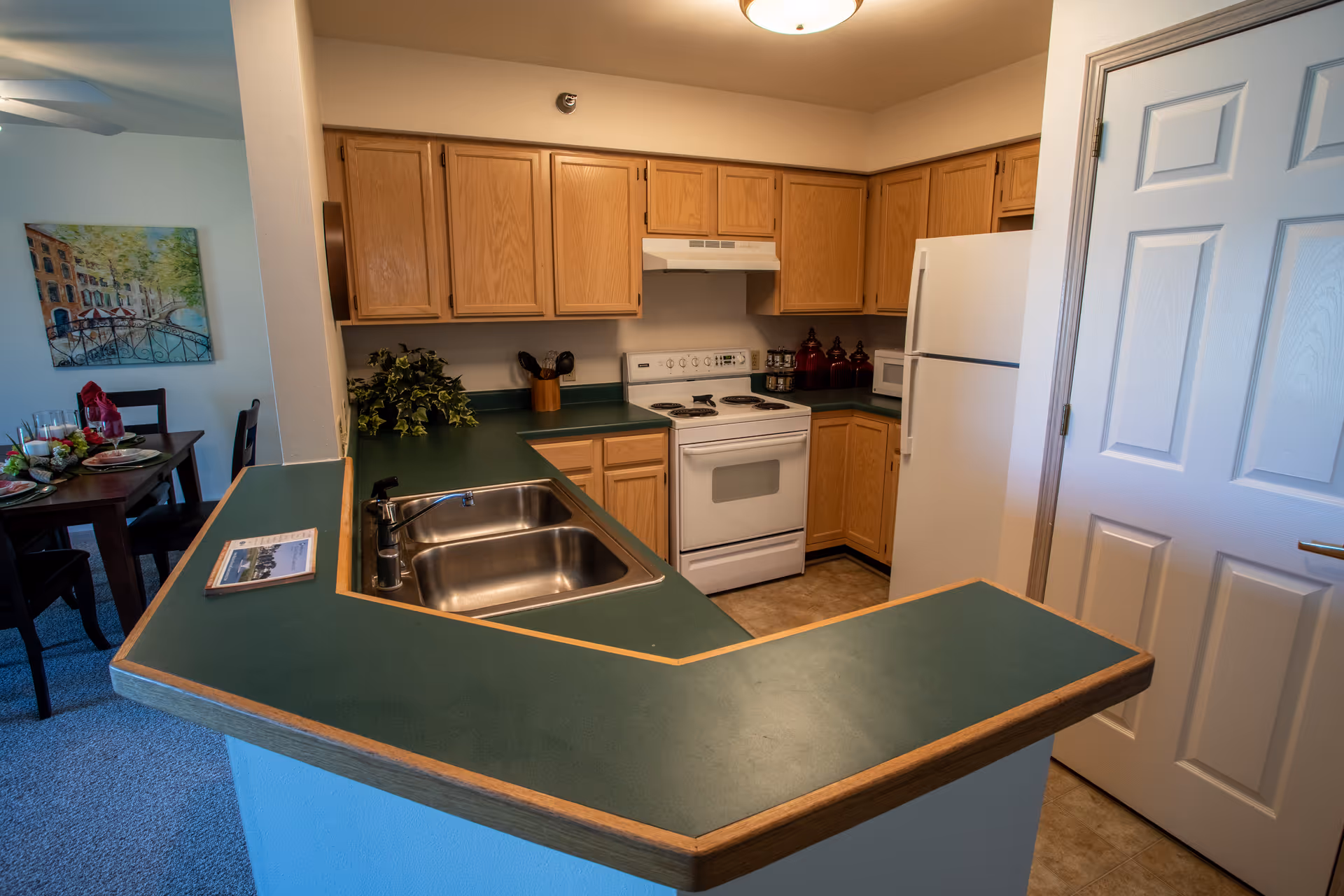 Small open kitchen with green laminate countertops, oak cabinets, a white stove and refrigerator, and a double sink in a peninsula overlooking a dining area.