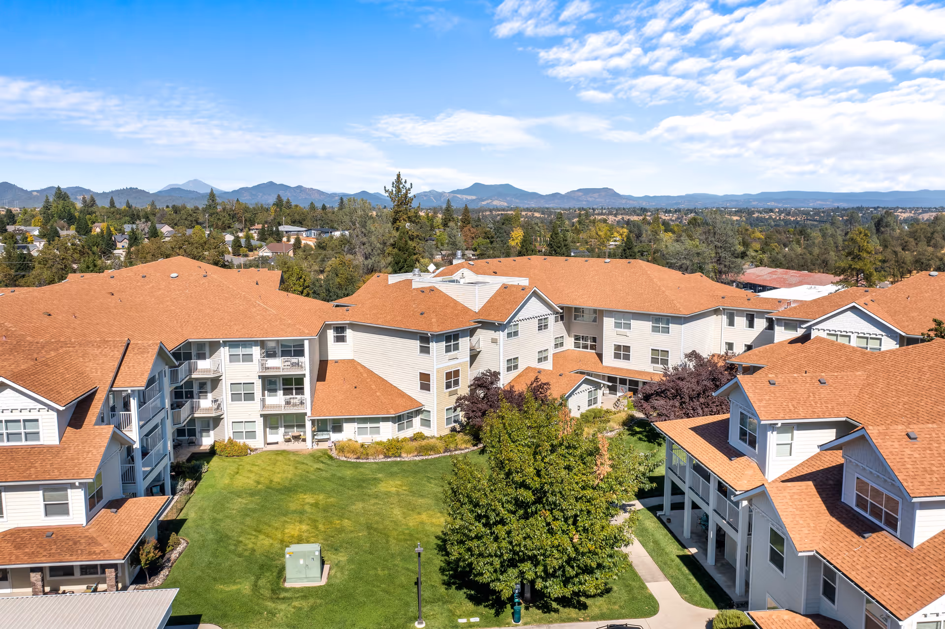 Aerial view of Shasta Estates Senior Living facility showing multiple connected buildings with red roofs surrounding a green lawn and trees, with mountains and a partly cloudy sky in the background.