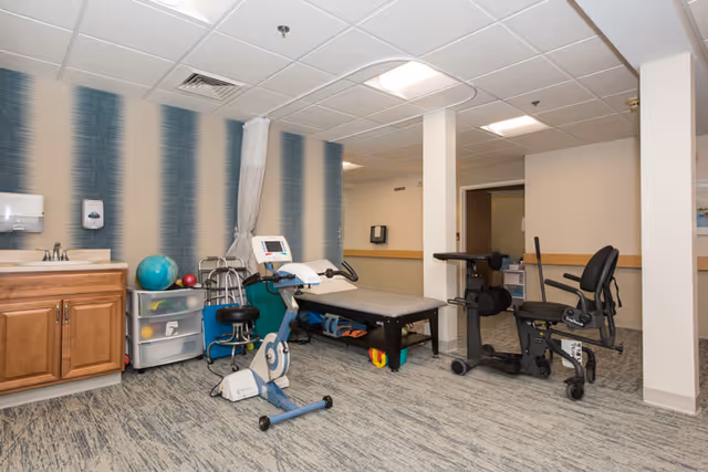 A rehabilitation room with exercise equipment including a stationary bike, a therapy table, and a leg exercise machine. The room has a patterned carpet, a sink with wooden cabinets, a small storage drawer unit, and a wall with vertical blue and white stripes. The ceiling has recessed lighting and a ventilation vent.