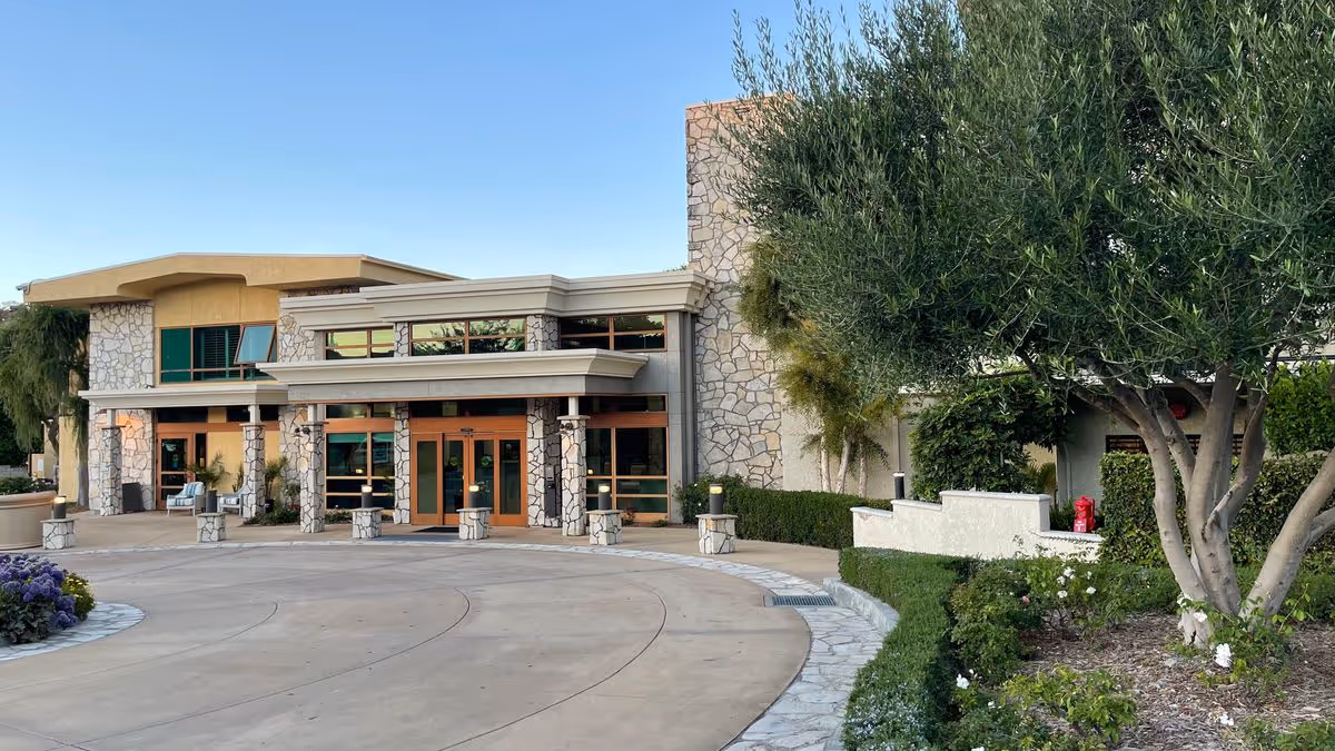 Front entrance of a stone-faced retirement community building with a circular driveway and landscaped trees and shrubs.