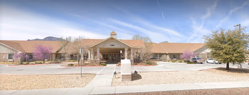 Front exterior view of Los Arcos del Norte Care Center building with a covered entrance, a driveway, several trees with purple blossoms, parked cars, and a clear blue sky.