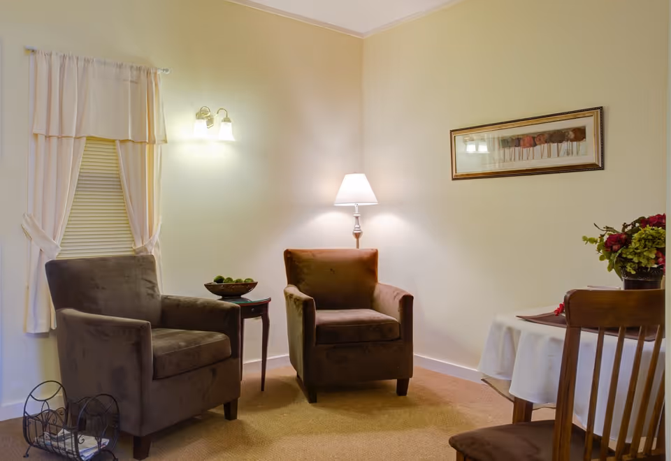 A cozy sitting area with two brown upholstered armchairs, a small wooden side table with a bowl of green decorative balls, a floor lamp, a framed artwork on the wall, and a small table with a white tablecloth and a floral arrangement. The room has beige walls and carpet, with a window covered by white curtains and blinds.
