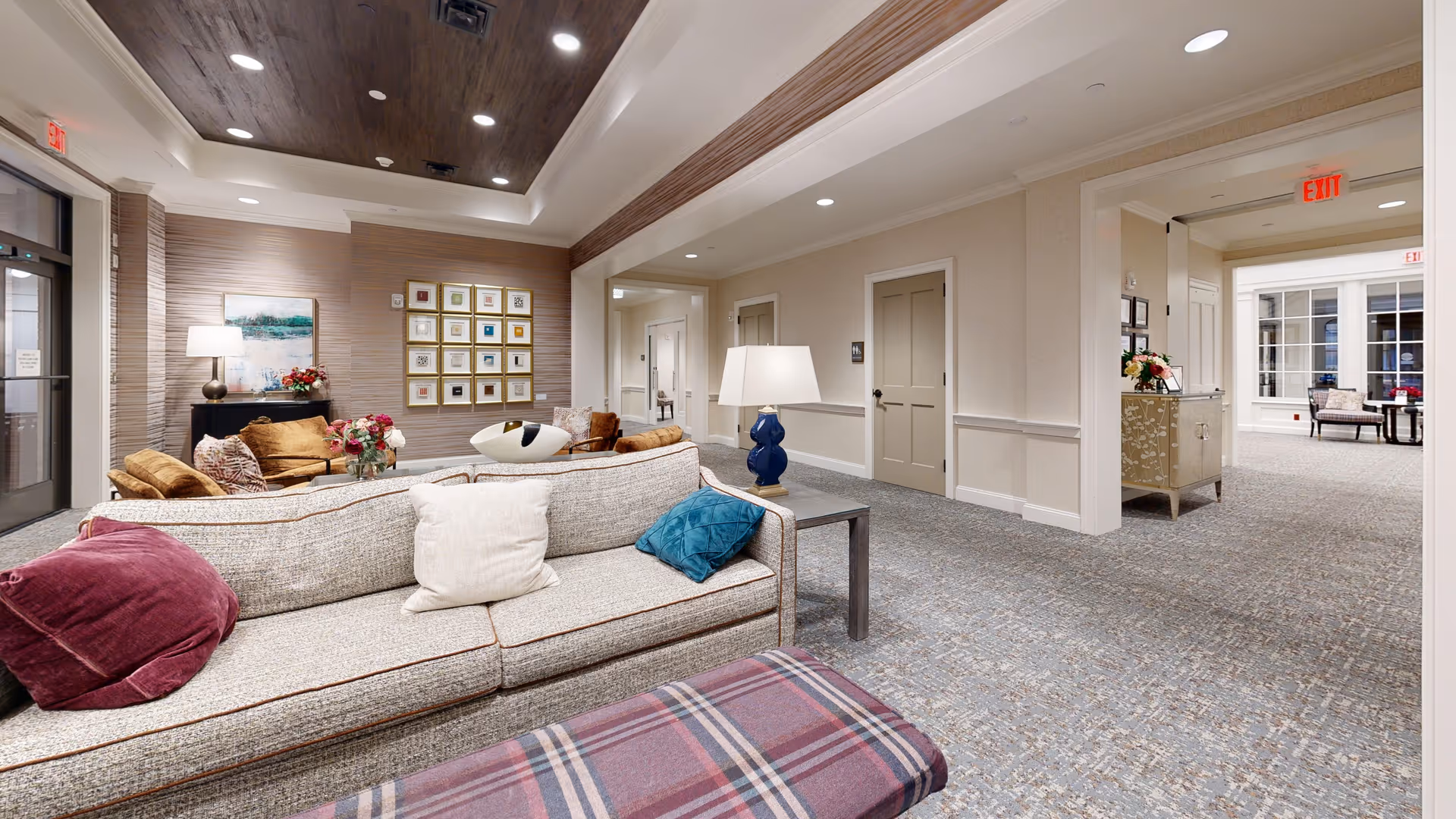A spacious and well-lit living room area in a senior living facility with a beige sofa adorned with colorful pillows, a plaid ottoman, a side table with a blue lamp, and additional seating with brown chairs. The walls are decorated with framed artwork and a painting, and the carpeted floor extends into a hallway with multiple doors and an exit sign.
