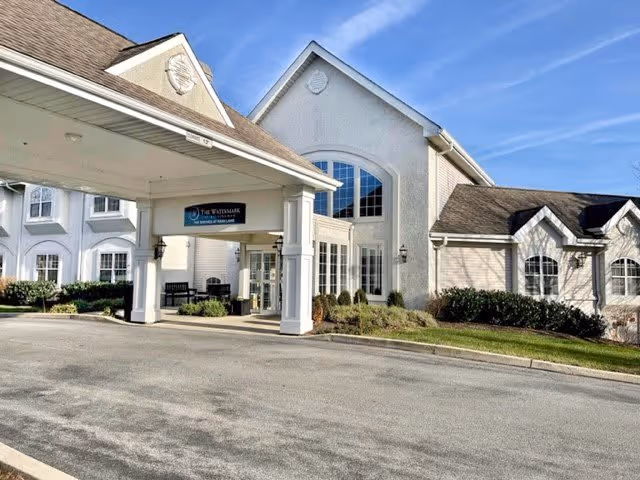 Exterior view of a senior living facility with a covered entrance, white walls, large windows, and a well-maintained lawn under a clear blue sky.
