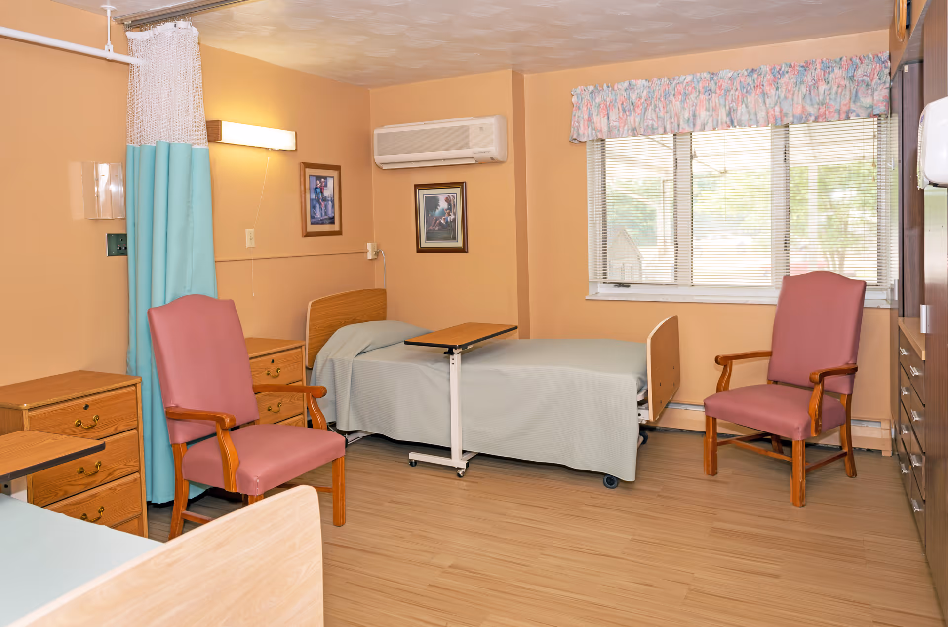 A senior living facility bedroom featuring two beds, an overbed table, bedside dressers, two pink armchairs, and a window.