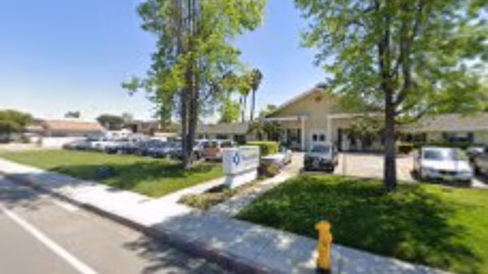 Front exterior of a single-story post-acute rehab building with a parking lot, grassy lawn, trees, and a sidewalk.