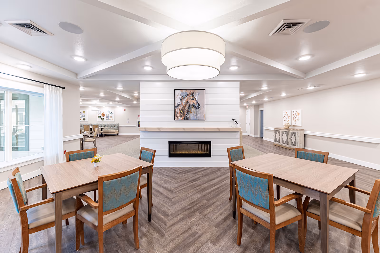 A bright and spacious senior living common area with two wooden tables, each surrounded by four chairs with blue patterned upholstery. A modern electric fireplace is centered on a white shiplap wall, above which hangs a painting of a horse. The room features wood-patterned flooring, a large circular ceiling light fixture, and large windows with white curtains allowing natural light to enter. Additional seating and decorative elements are visible in the background.