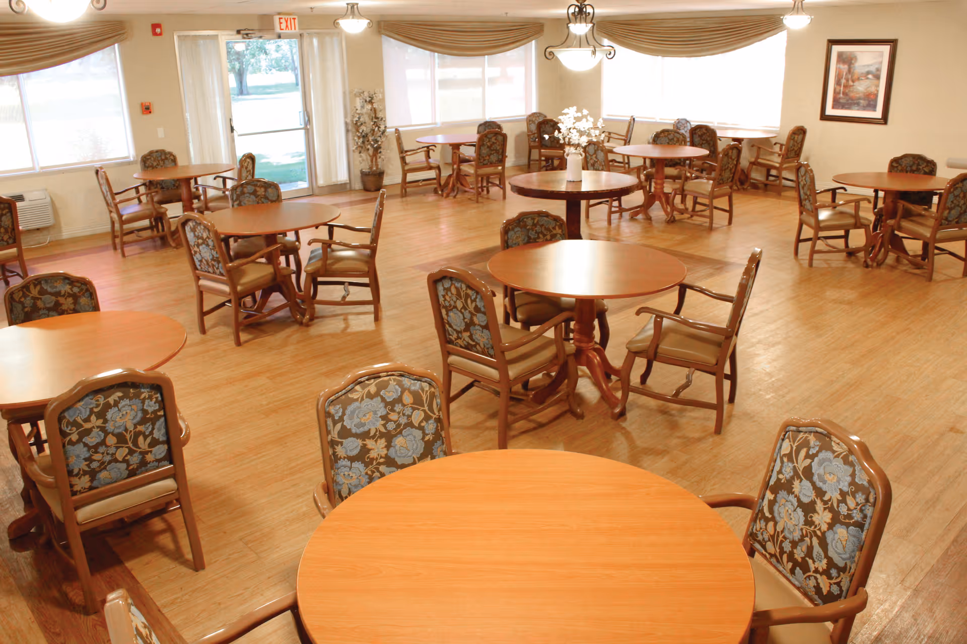 A spacious dining room with multiple round wooden tables and chairs with floral upholstery. The room has large windows with beige valances, wooden flooring, and a framed picture on the wall. There is a glass door leading outside and some decorative plants near the door.