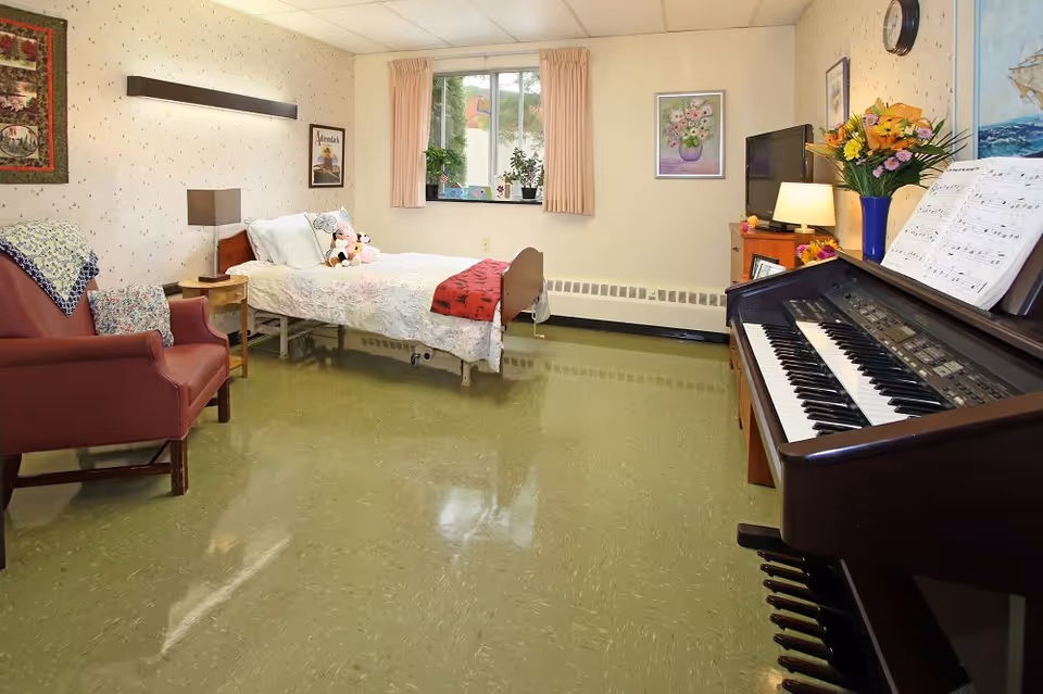 A cozy bedroom in a senior living facility featuring a single bed with white bedding and stuffed animals, a red armchair with a crocheted blanket, a wooden nightstand with a lamp, a window with beige curtains, a TV on a wooden dresser, a vase of flowers, and an electric organ with sheet music.