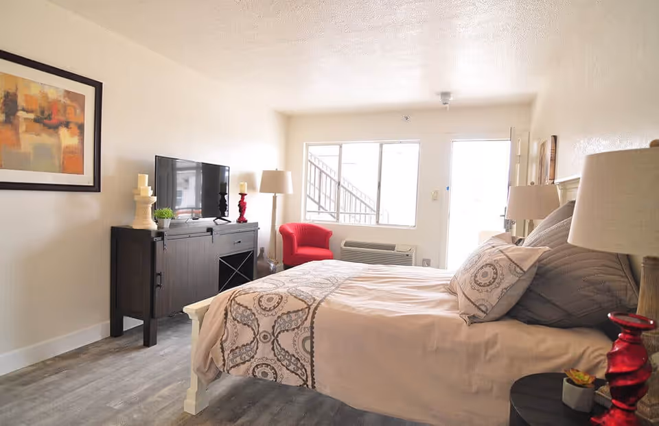 A bright bedroom with a large bed covered in beige and patterned bedding. There are two pillows on the bed, a bedside table with a lamp and a red decorative item, and a dark wooden cabinet with a flat-screen TV and candles on top. A red armchair is placed near a window and an air conditioning unit below it. The room has light-colored walls and wood flooring, with a framed abstract painting on one wall.