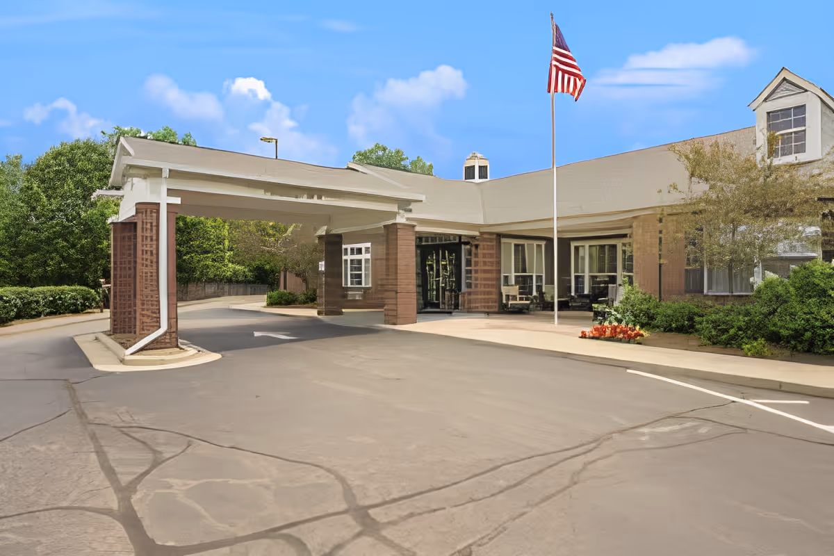 Exterior view of the entrance to Rittenhouse Village At Northside, featuring a covered driveway supported by brick columns, an American flag on a flagpole, and surrounding greenery under a blue sky with some clouds.