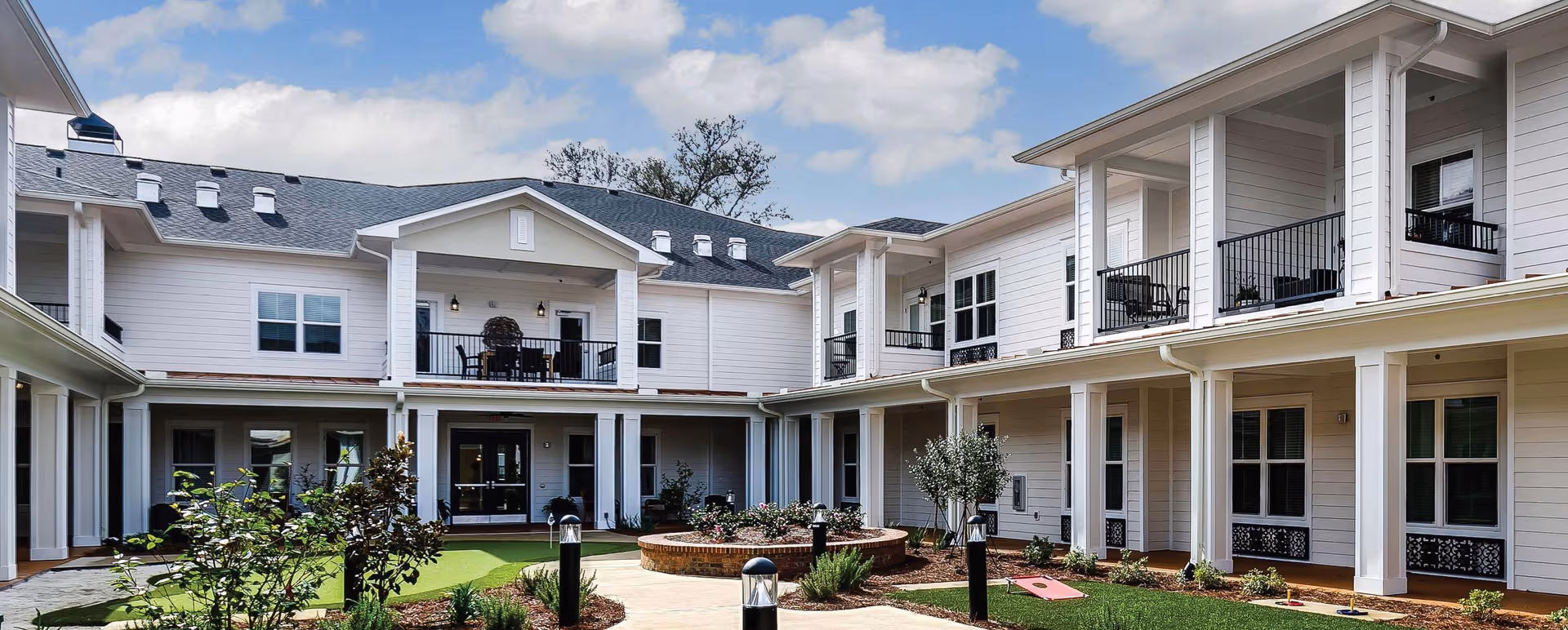 Outdoor courtyard area of The Blake at New Braunfels facility featuring a two-story white building with balconies, a landscaped garden with plants and flowers, a circular brick planter, and a putting green with golf holes and a cornhole game set up under a partly cloudy sky.