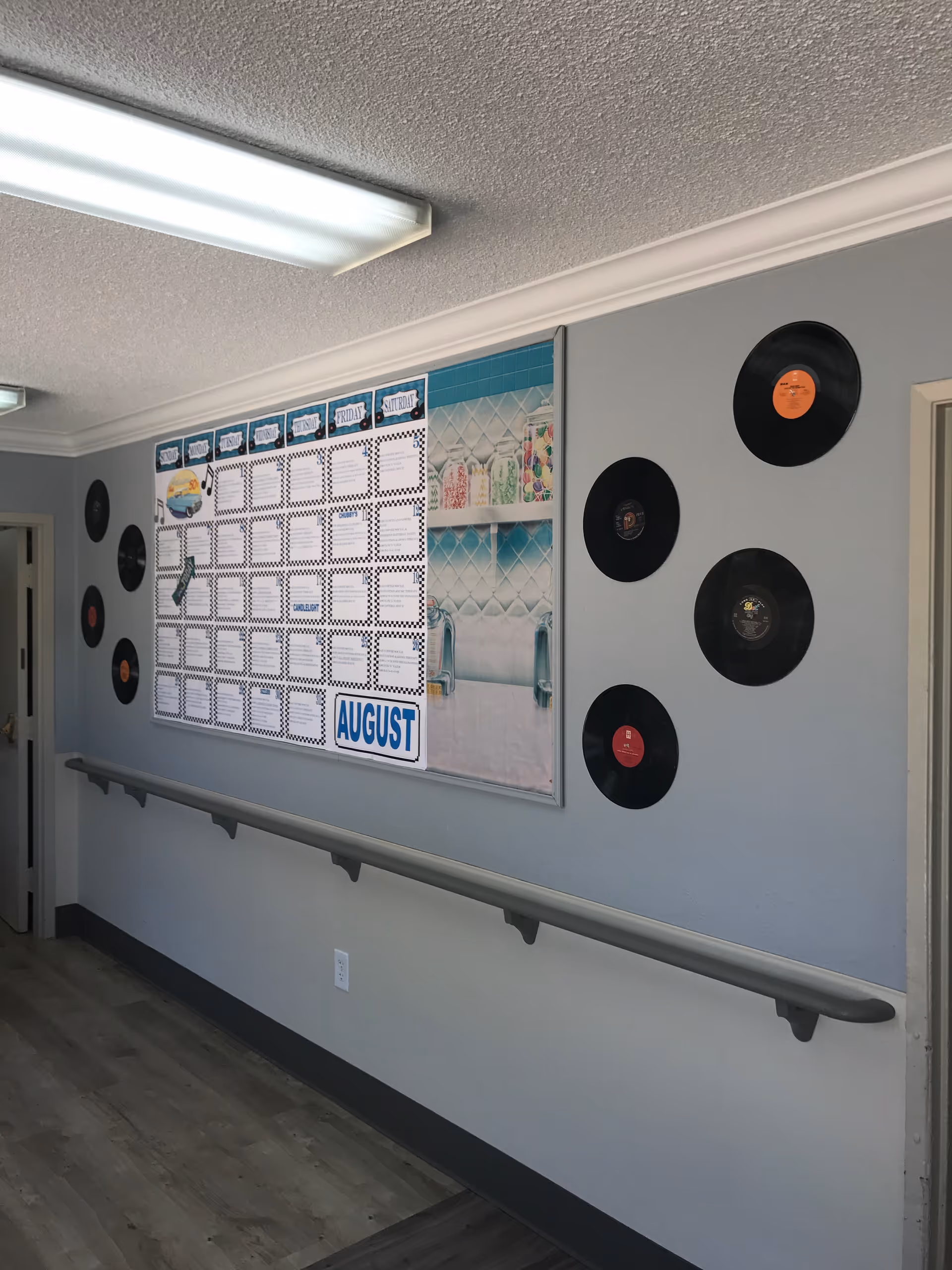 A hallway with a handrail along the wall, decorated with a large monthly calendar for August and several vinyl records mounted on the wall. The floor is wood, and the walls are painted light gray with a darker gray baseboard. There are ceiling lights and a doorway visible at the end of the hallway.
