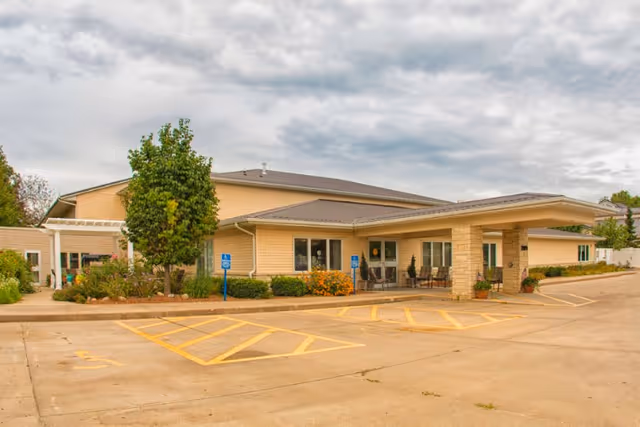Exterior view of a single-story assisted living facility building with beige siding, a covered entrance supported by stone pillars, several windows, and a parking area with marked handicap spaces in front. There are some shrubs and a tree near the entrance, and the sky is cloudy.
