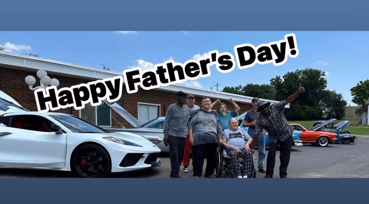 A group of seven men, including one in a wheelchair, posing happily in front of several classic and modern cars with their hoods open in a parking lot outside a brick building under a blue sky. The text 'Happy Father's Day!' is prominently displayed above them.
