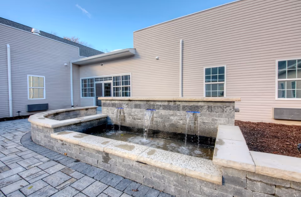 Outdoor courtyard area at Meadows Assisted Living and Care Campus featuring a stone water fountain with three spouts flowing water into a rectangular basin, surrounded by paved stone flooring and beige siding building walls with windows in the background under a clear blue sky.