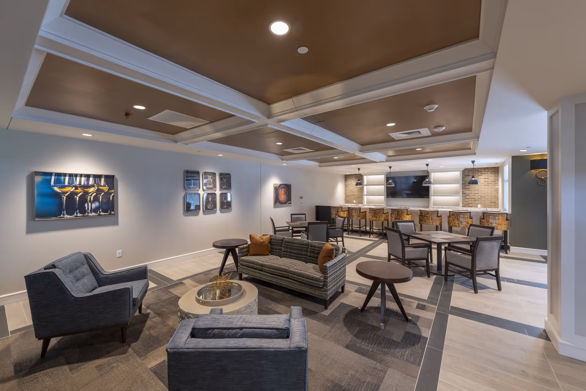 A modern lounge area with a plaid sofa, two gray armchairs, round side tables, and a circular coffee table. The room features a coffered ceiling with recessed lighting, artwork on the walls, and a bar area with high chairs and pendant lights in the background.