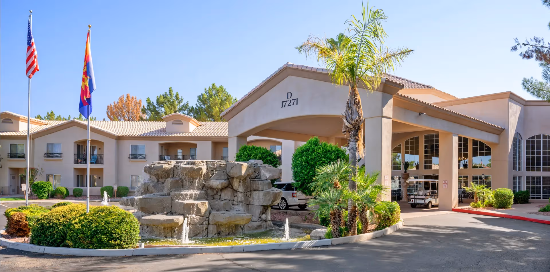 Exterior view of The Montecito Senior Living facility featuring a beige building with a tiled roof, a covered entrance with the number D 17271, a rock fountain with water flowing, two flagpoles with the American and Arizona state flags, and surrounding greenery including palm trees and bushes under a clear blue sky.