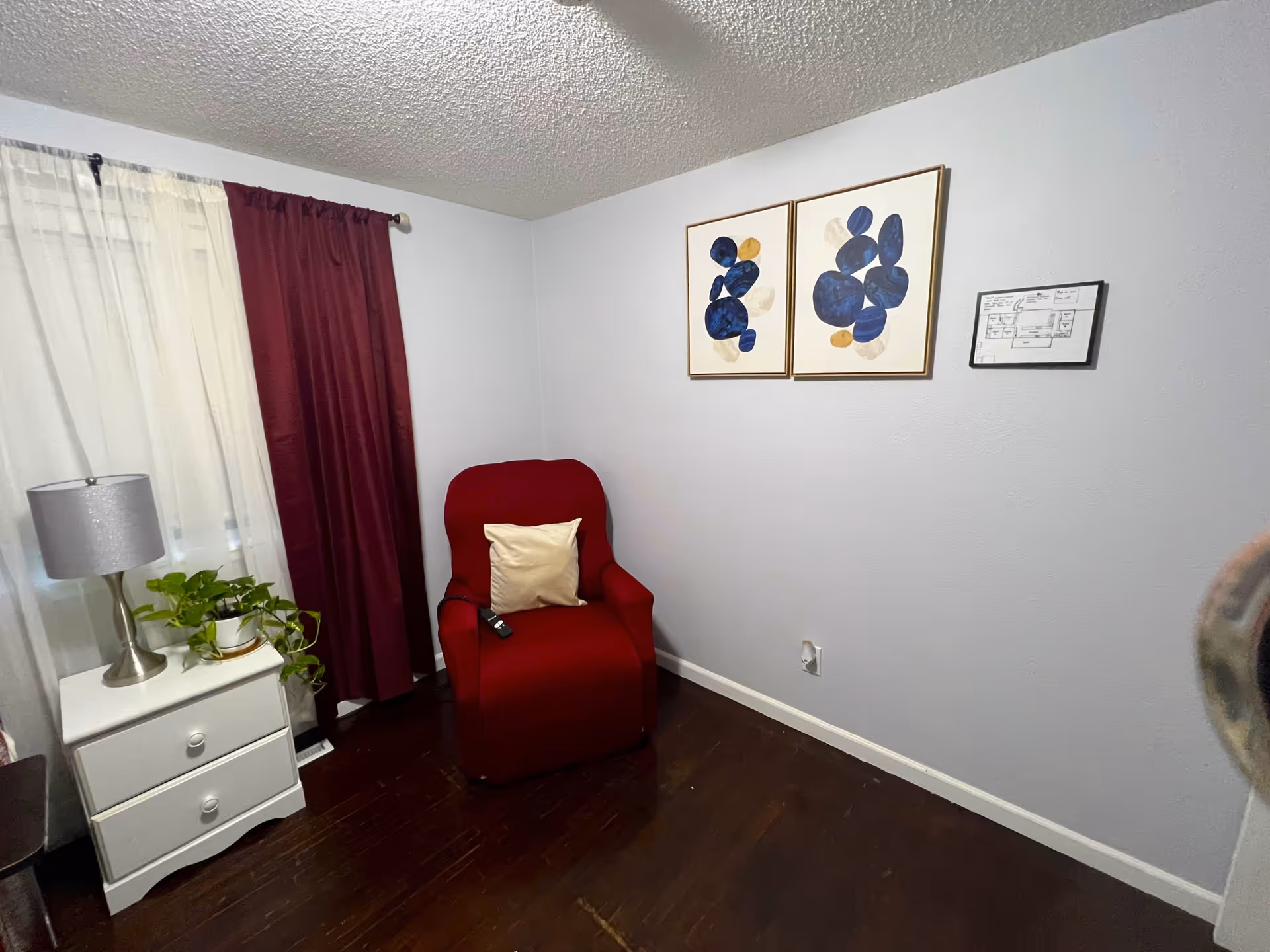 A cozy corner of a room with a red armchair featuring a beige pillow and a remote control on it. Next to the chair is a white nightstand with two drawers, a silver lamp, and a green potted plant. The wall behind the chair is light gray and decorated with two abstract blue and gold framed artworks and a small framed floor plan. The window has white and dark red curtains, and the floor is dark wood.
