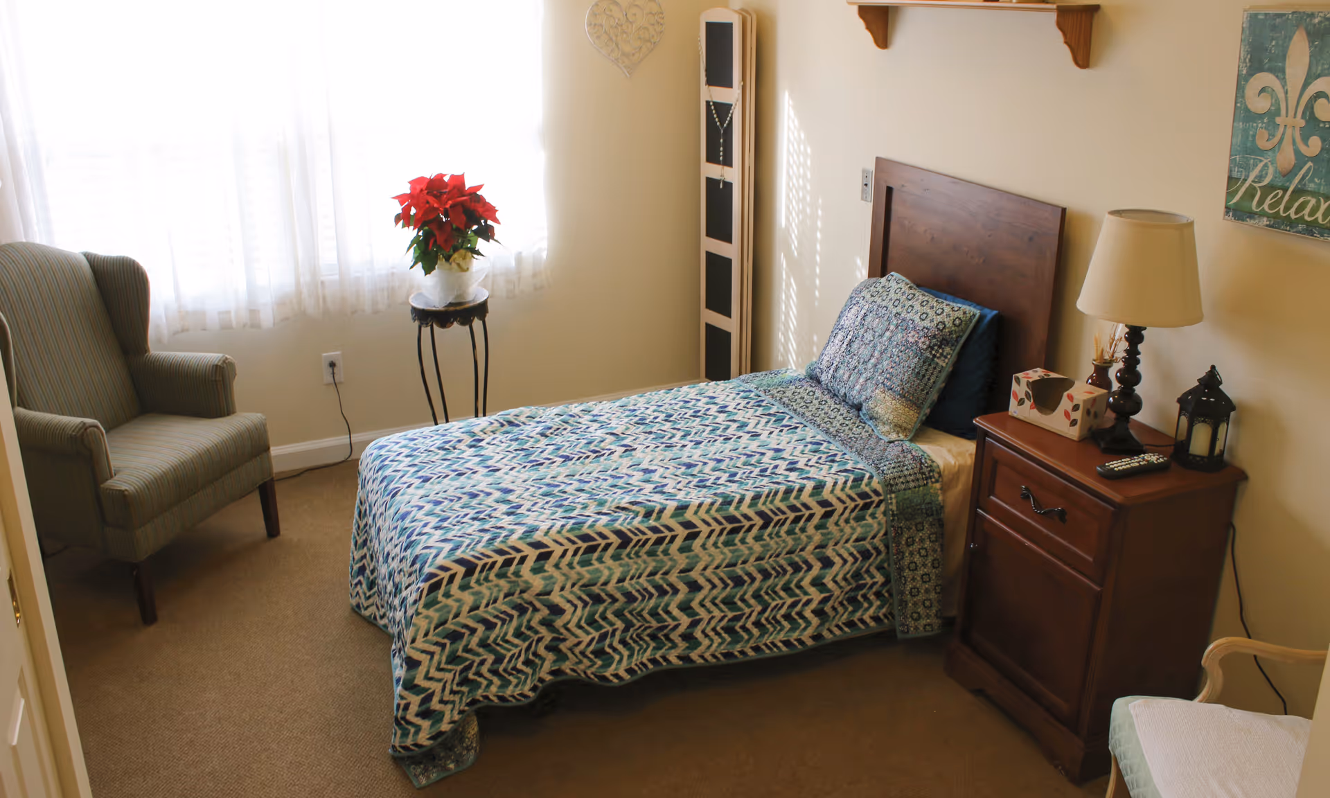A tidy single-occupancy bedroom with a bed, nightstand, lamp and armchair beside a curtained window.