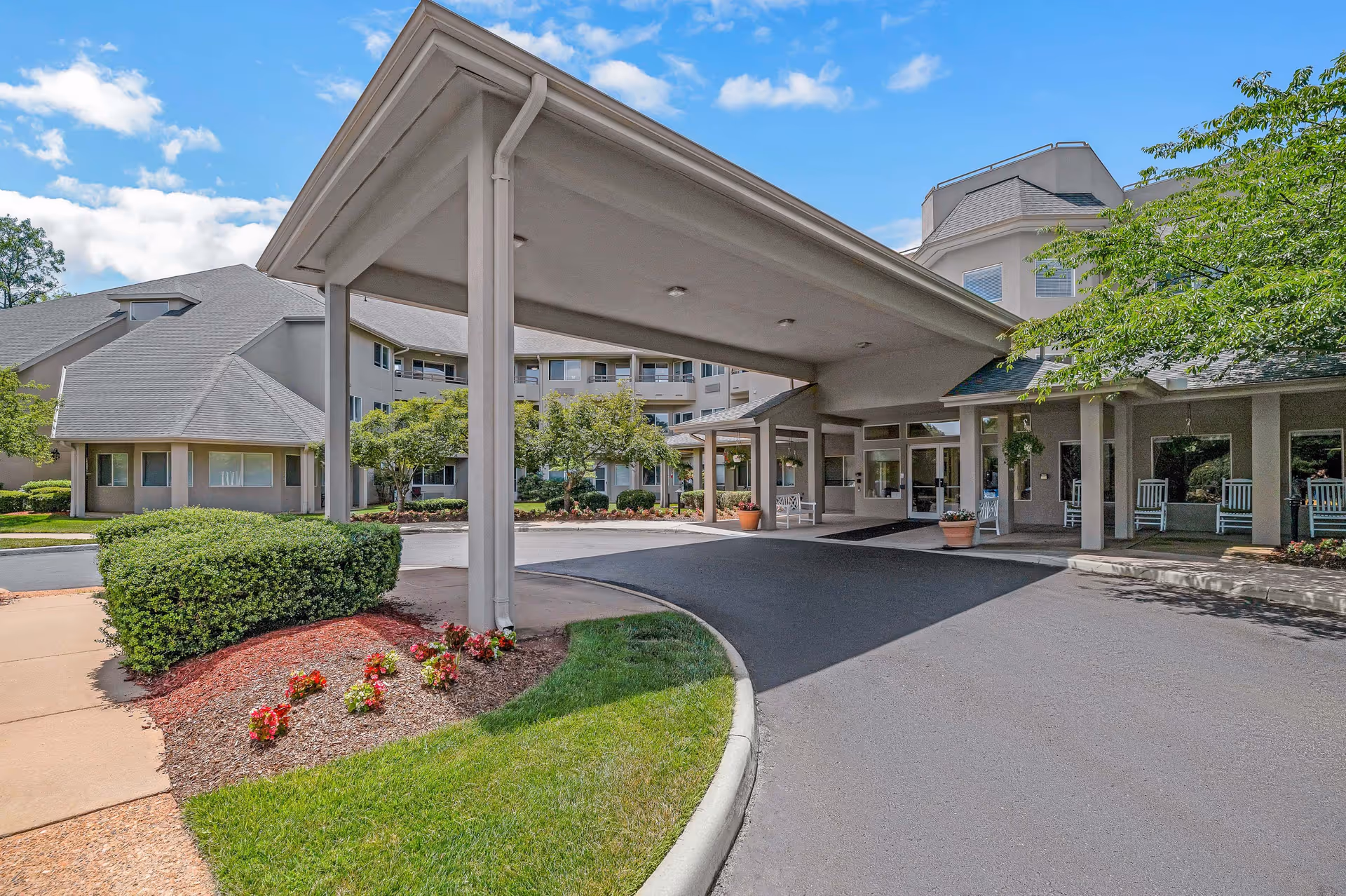 Covered porte-cochere entrance of a multi-story senior living building with driveway, landscaping, and rocking chairs on the porch.