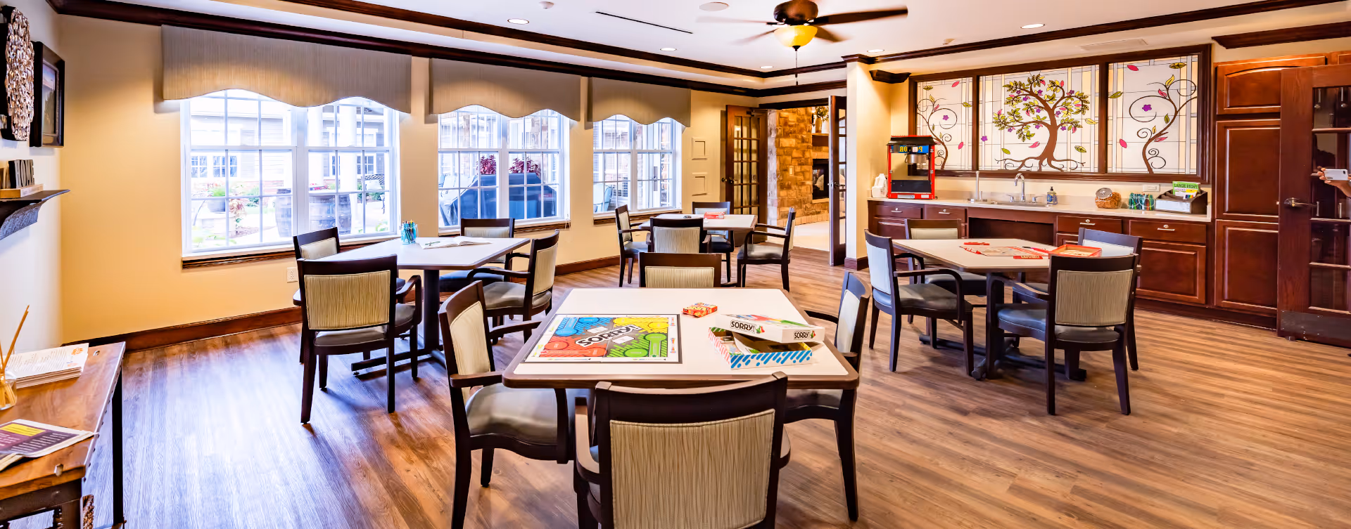 A bright and spacious common room with multiple tables and chairs arranged for group activities. The room features large windows with valances, wooden flooring, and a ceiling fan with a light. On the tables, there are board games like Sorry! and other game pieces. The back wall has a countertop with a sink, cabinets, and decorative stained glass panels with a tree and floral design. A popcorn machine is also visible on the counter.