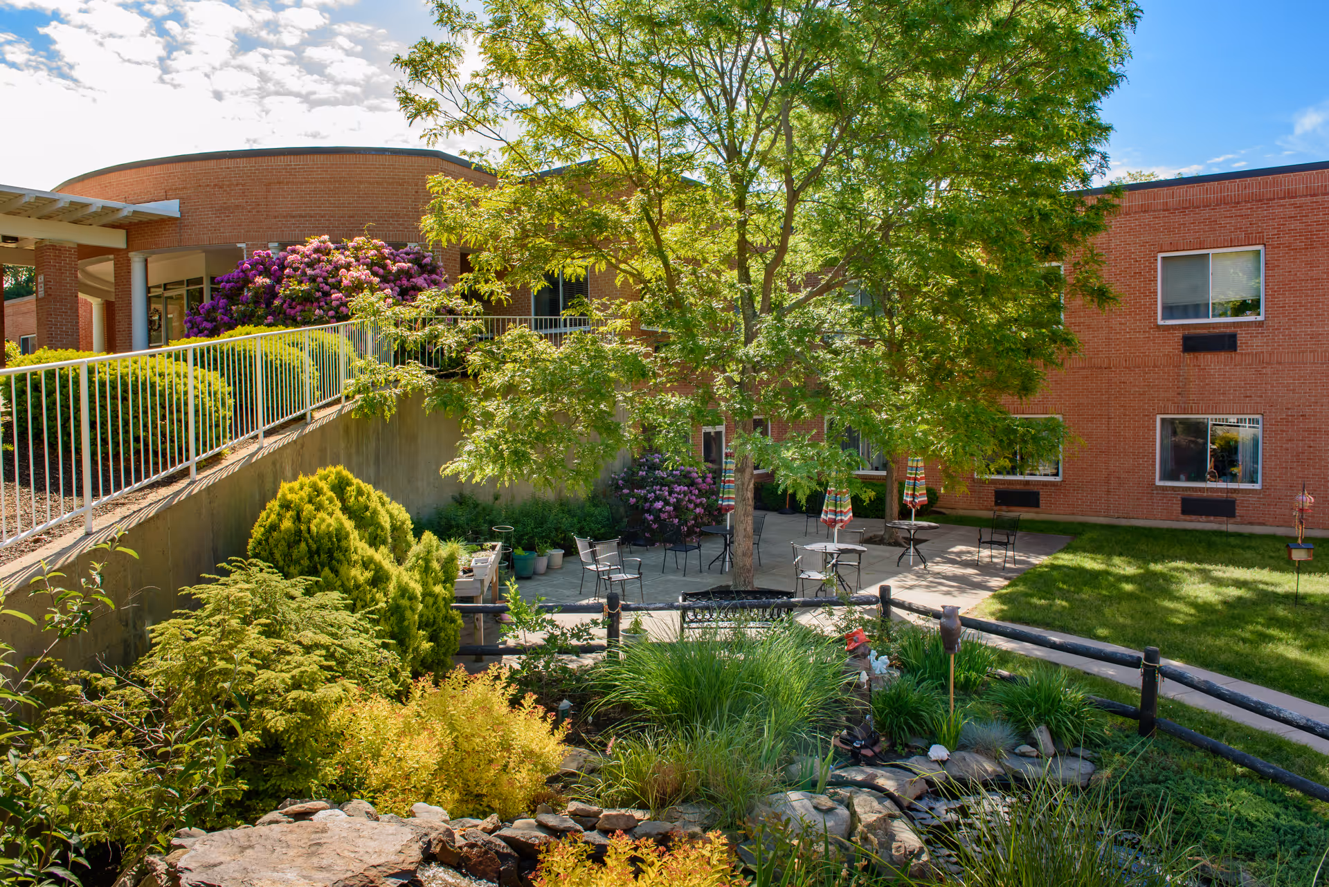 Outdoor courtyard area at a senior living facility with a large tree providing shade over several tables and chairs with umbrellas. Surrounding the courtyard are green bushes, flowering plants, and a small rock garden. The building's brick exterior and windows are visible in the background under a partly cloudy blue sky.
