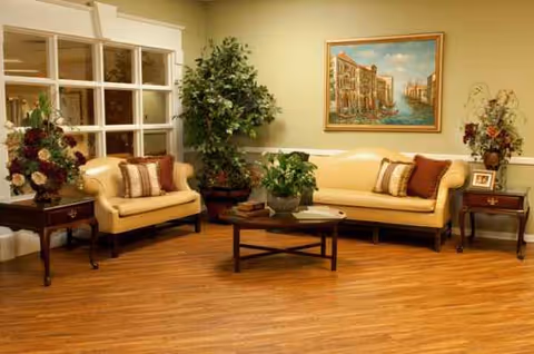 Bright senior living facility lounge with beige couches, plants, and a central coffee table.