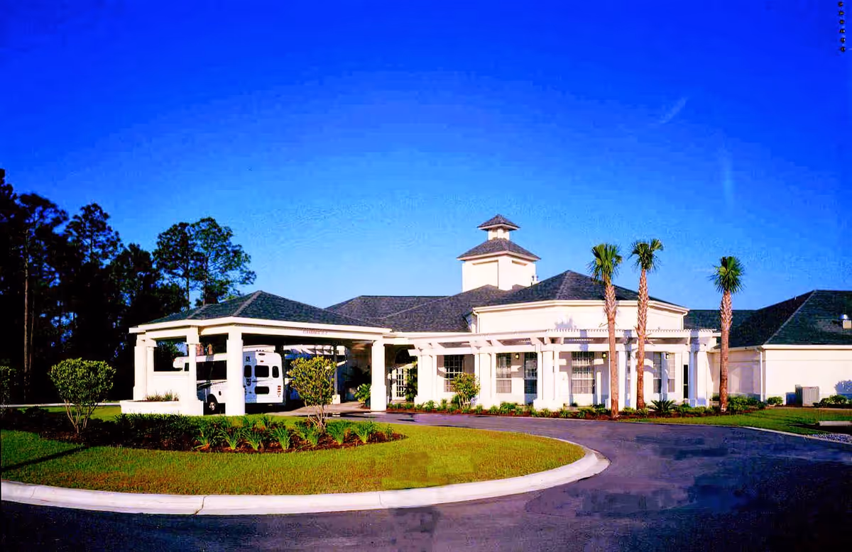 Exterior view of a single-story building with a covered entrance and a circular driveway. The building is surrounded by well-maintained landscaping including palm trees, bushes, and grass. A white van is parked under the covered entrance. The sky is clear and blue.