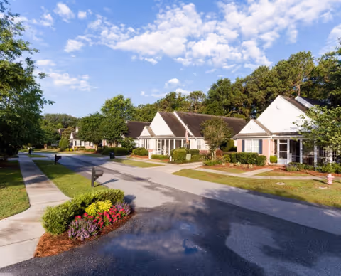 A quiet residential street in a senior living community with single-story houses, well-maintained lawns, bushes, and trees under a partly cloudy blue sky.
