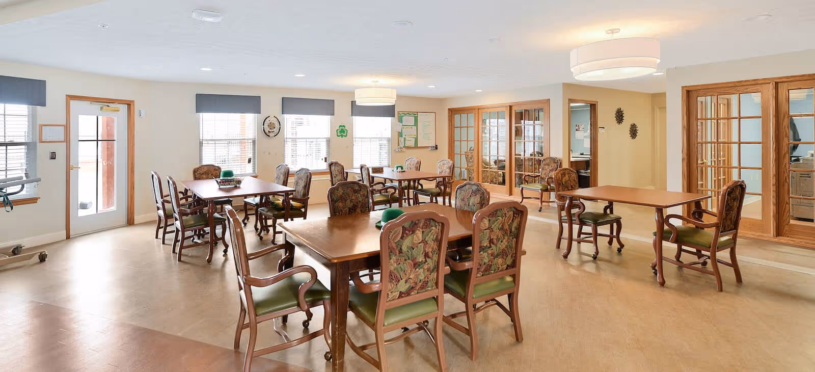 Bright communal dining/activity room with multiple wooden tables and upholstered chairs, large windows, and glass French doors.