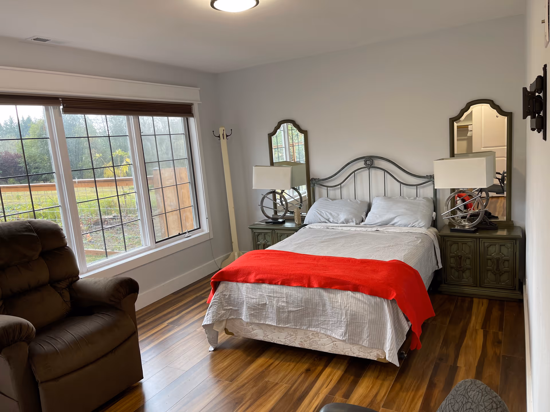 A bedroom with a double bed covered in white bedding and a red blanket at the foot. The bed has a metal headboard and is flanked by two green nightstands, each with a lamp and a mirror behind it. To the left, there is a large window with a view of greenery outside and a brown recliner chair. The floor is wooden, and the walls are painted white.