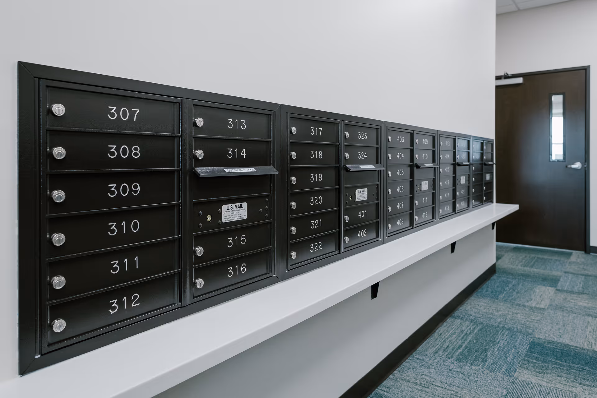 A row of black mailboxes mounted on a white wall inside a building, each labeled with numbers such as 307 to 324 and 401 to 412. The mailboxes have small locks and some have mail slots. The floor is carpeted in a blue and green pattern, and there is a dark door with a narrow window at the end of the hallway.