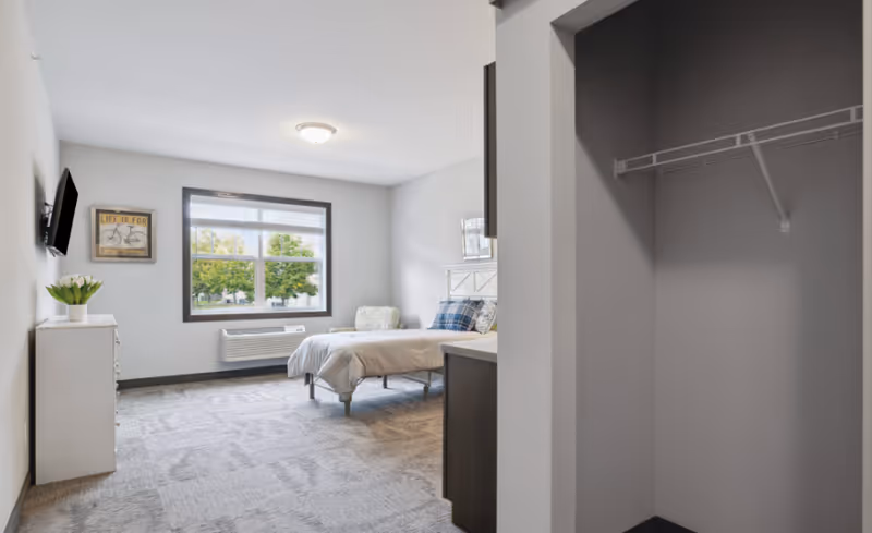 A bright and clean senior living bedroom with a bed, a chair, a dresser with a vase of flowers, a wall-mounted TV, and a window showing green trees outside. There is an open closet with a wire shelf on the right side.