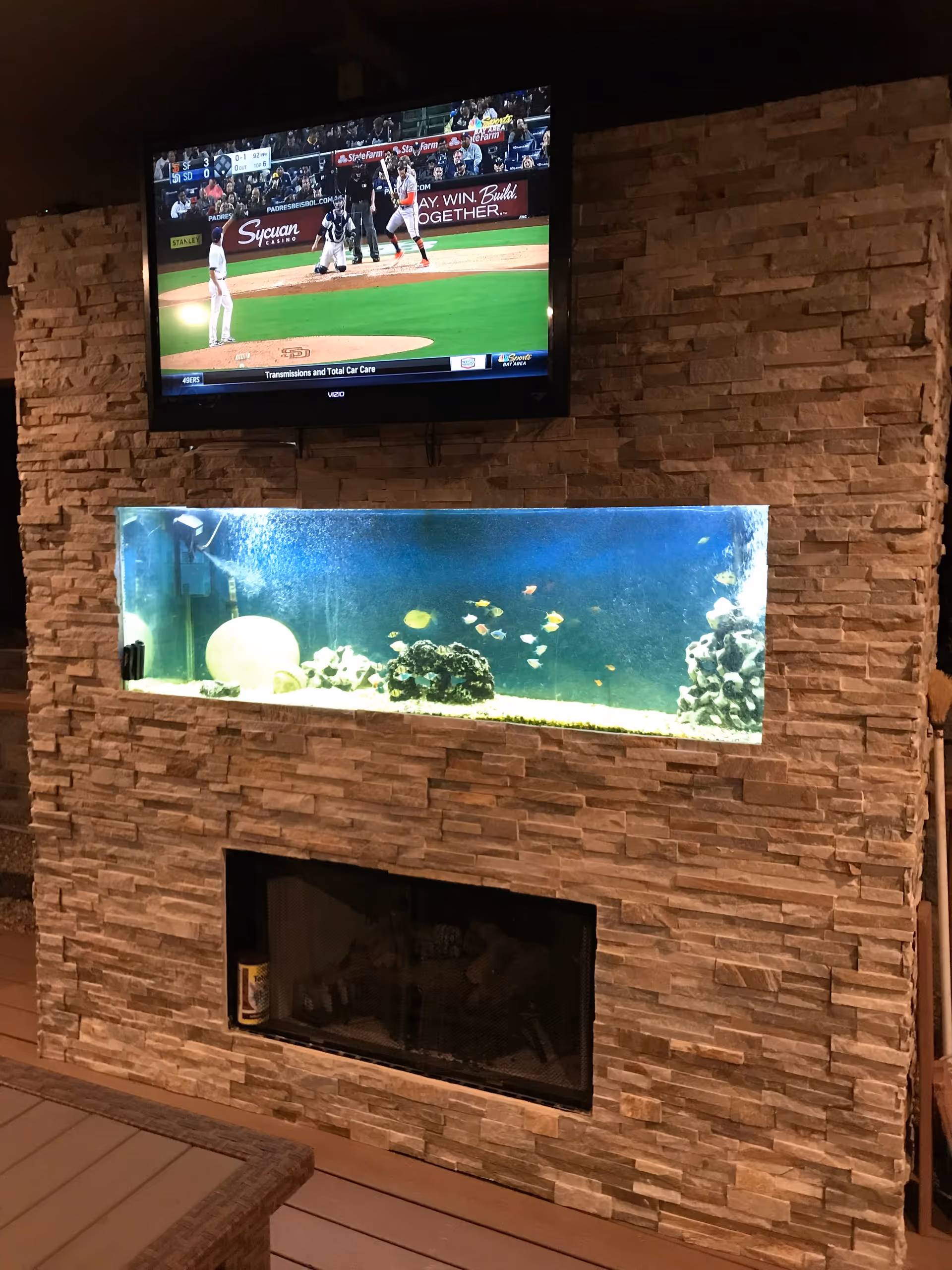 A stone wall with a built-in aquarium filled with small colorful fish. Above the aquarium, a flat-screen TV is mounted on the wall showing a baseball game. Below the aquarium, there is a fireplace with a glass cover.