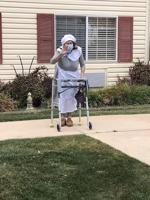 An elderly woman wearing a white bonnet, apron, and face mask is standing outdoors on a sidewalk using a walker. She is waving with one hand. Behind her is a beige building with red shutters and a window.