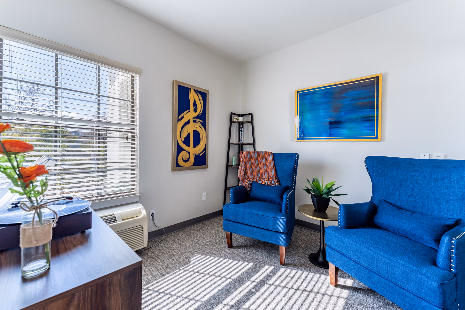 A bright room with two blue upholstered armchairs, one draped with an orange throw blanket, a small round side table with a green plant, a tall black shelving unit in the corner, and two framed artworks on the walls. Sunlight streams through a window with white blinds, casting shadows on the carpeted floor. A wooden cabinet with a glass vase holding orange flowers is partially visible in the foreground.
