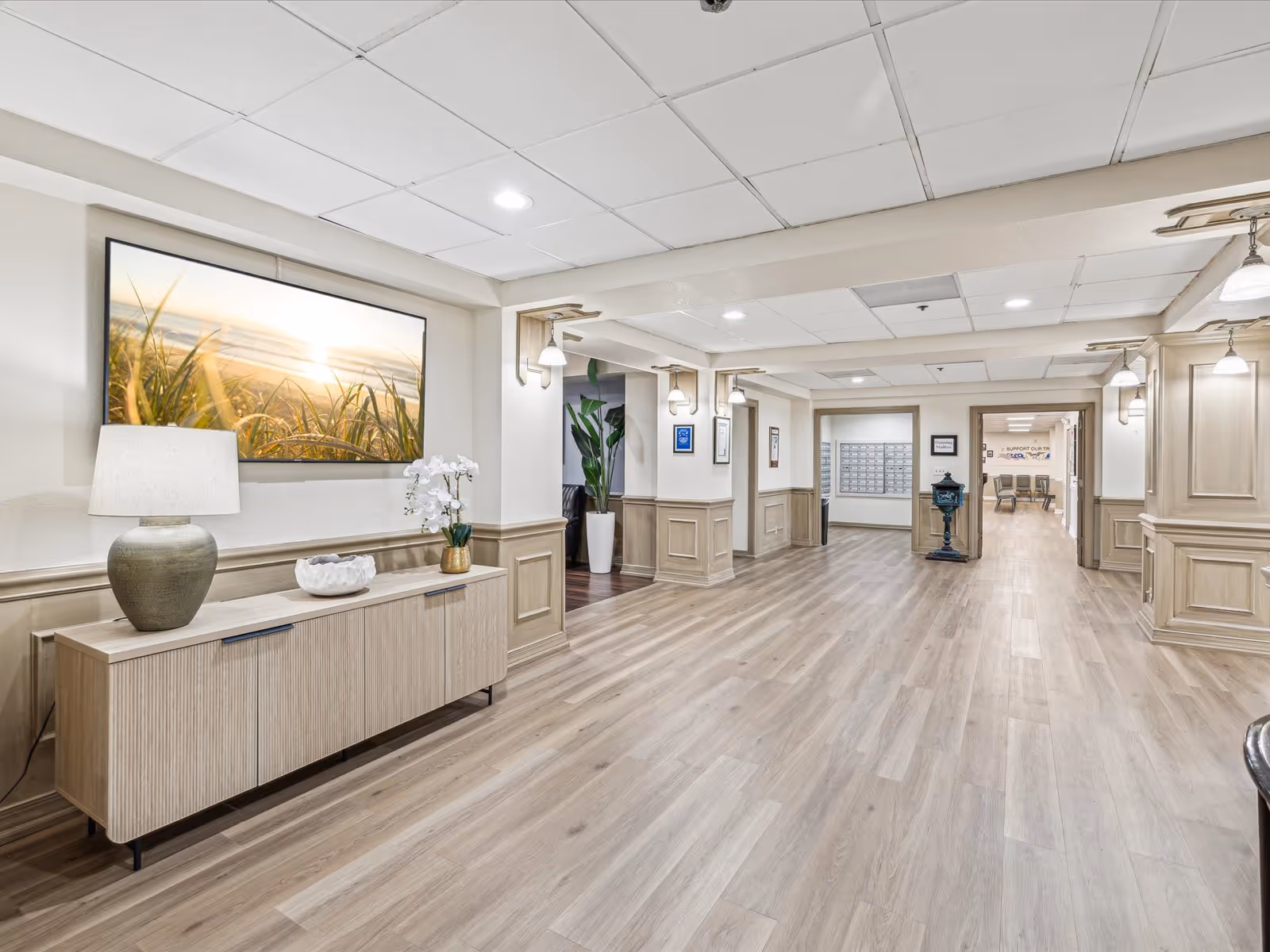 A spacious and well-lit hallway in a senior living facility with light wood flooring and beige wainscoting on the walls. On the left side, there is a modern sideboard with a lamp, a decorative bowl, and a white orchid plant. Above the sideboard is a large framed picture of a beach scene with grass and a sunset. The hallway leads to a room with mailboxes and another room with chairs visible in the background. The ceiling has recessed lighting and hanging light fixtures.