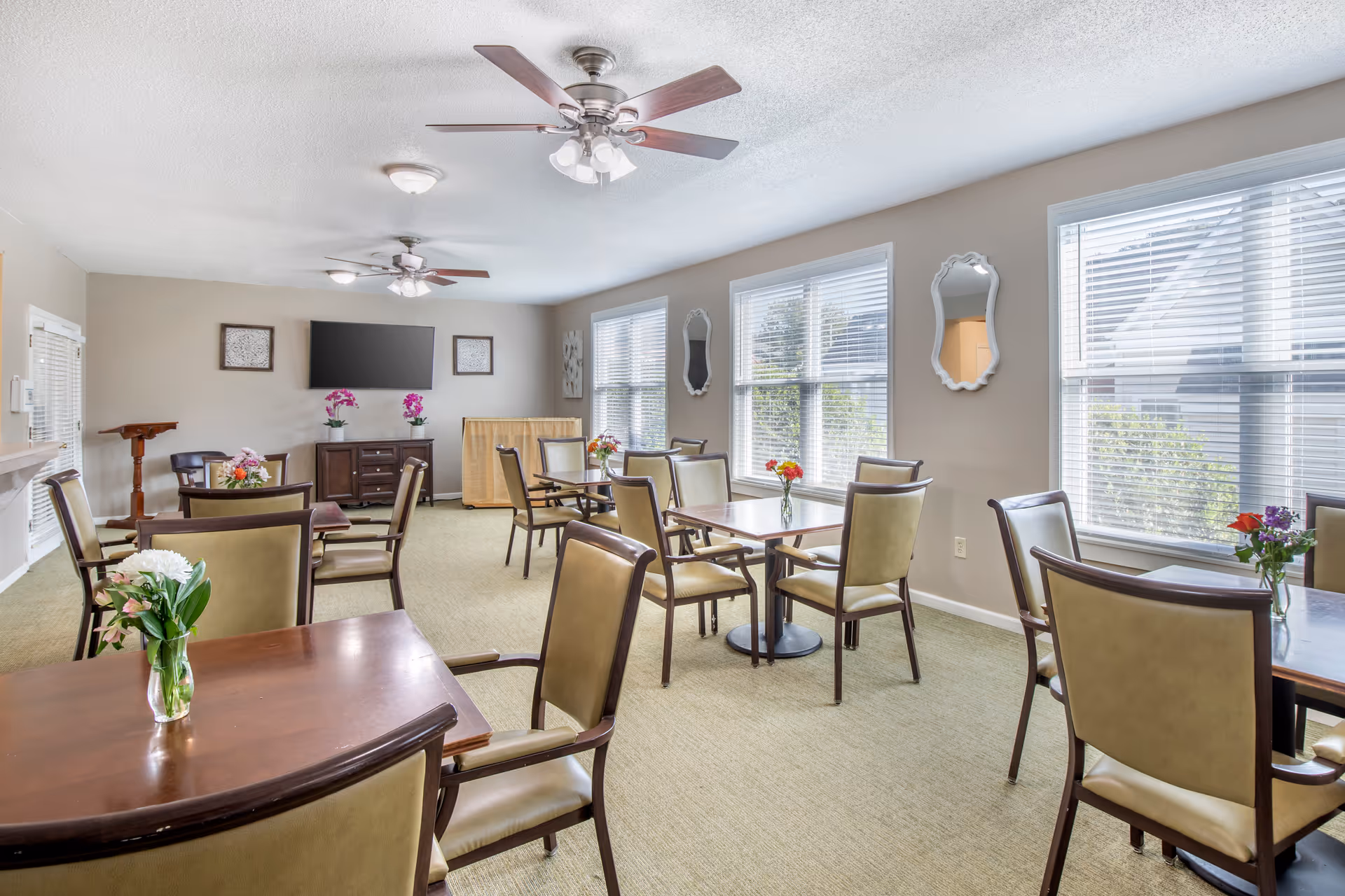 A bright dining room with several wooden tables and beige cushioned chairs arranged neatly. Each table has a small vase with colorful flowers. The room has large windows with white blinds letting in natural light, two ceiling fans with lights, a flat-screen TV mounted on the far wall, and decorative mirrors and artwork on the walls.