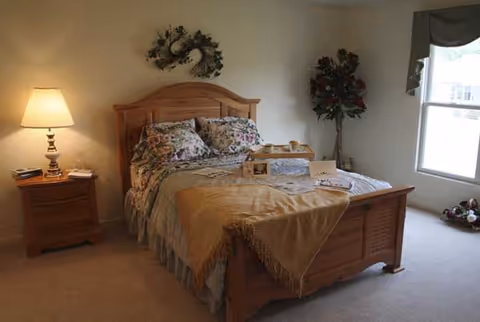 A cozy bedroom with a wooden bed frame and floral bedding. There is a nightstand with a lamp and books on the left side of the bed. A decorative wreath hangs on the wall above the bed, and a potted plant is placed near a window with curtains on the right side. The room is carpeted and well-lit with natural light coming through the window.
