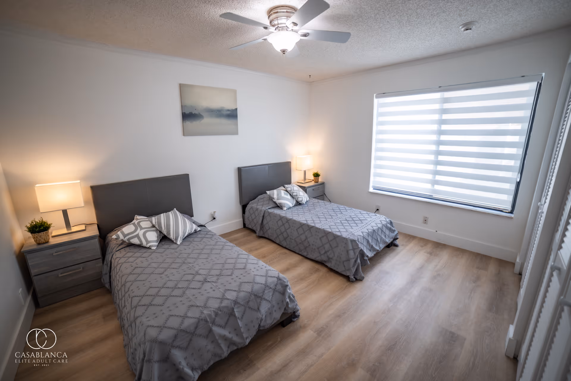 A clean and modern bedroom with two single beds, each with gray headboards and matching gray patterned bedspreads. Each bed has two decorative pillows. There are two nightstands, each with a lamp and a small potted plant. A large window with horizontal blinds allows natural light into the room. A ceiling fan with a light fixture is mounted on the ceiling. A framed picture hangs on the wall above the beds.